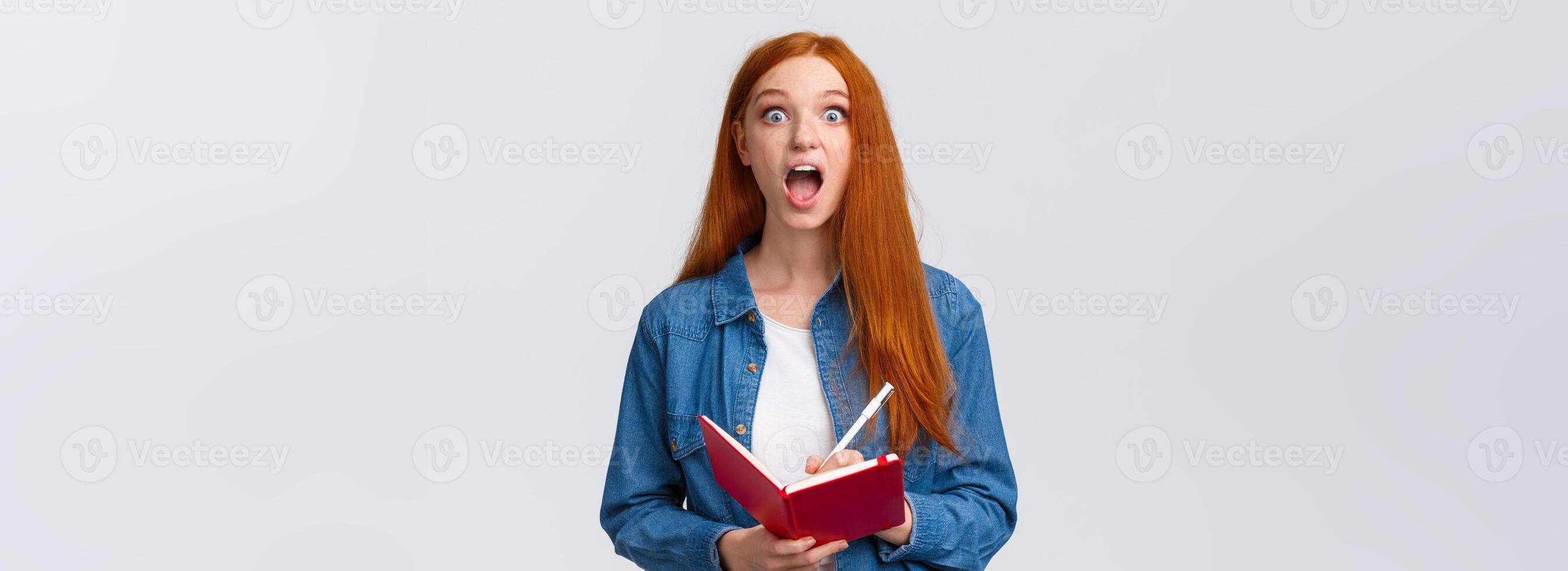 Amused and excited, astonished redhead girl fascinated with amazing lecture giving speech, writing down useful notes, holding notebook and staring thrilled camera, white background photo