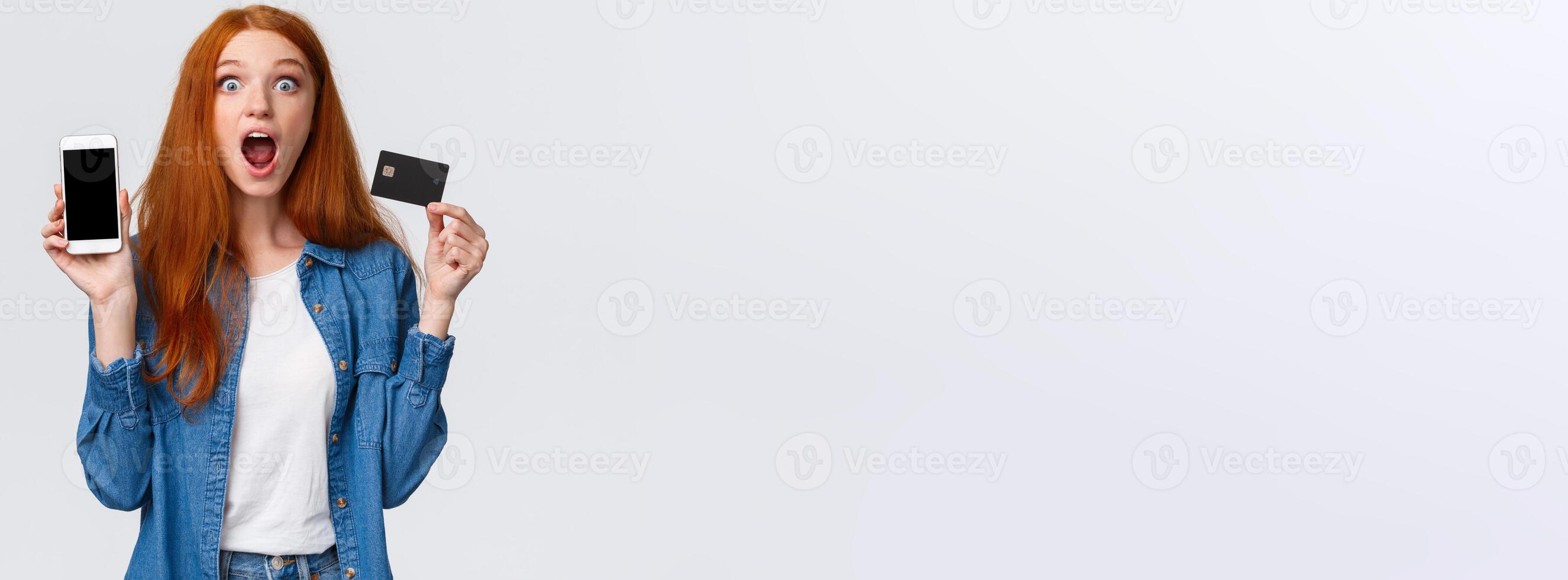 Waist-up portrait excited, astonished and thrilled redhead female student holding credit card, showing smartphone display, application, drop jaw impressed, looking camera astounded, white background photo