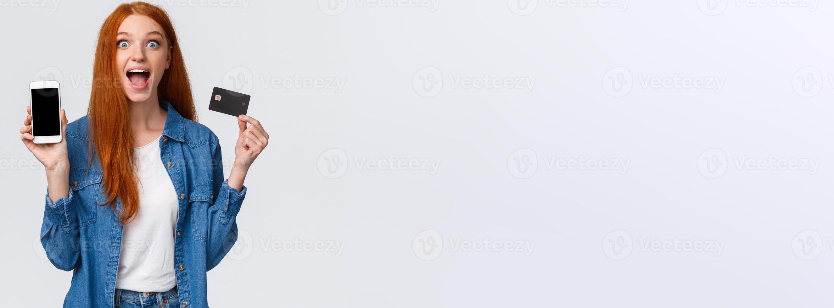 Amazed, happy and surpirsed, cheerful caucasian redhead woman showing credit card and smartphone display, app on mobile screen, scream amused standing white background photo
