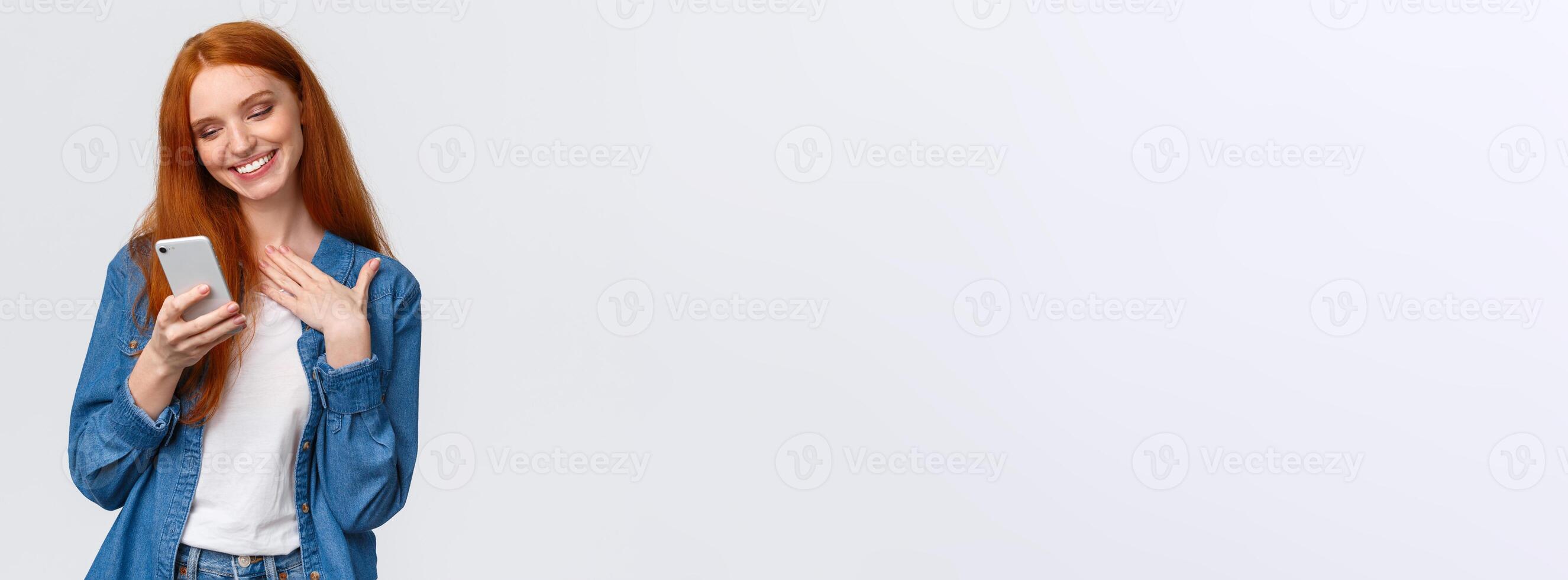 Oh so cute. Touched and pleased alluring redhead girlfriend receive love emoji, lovely message, texting, chatting boyfriend, holding smartphone, touch chest and smiling mobile display photo