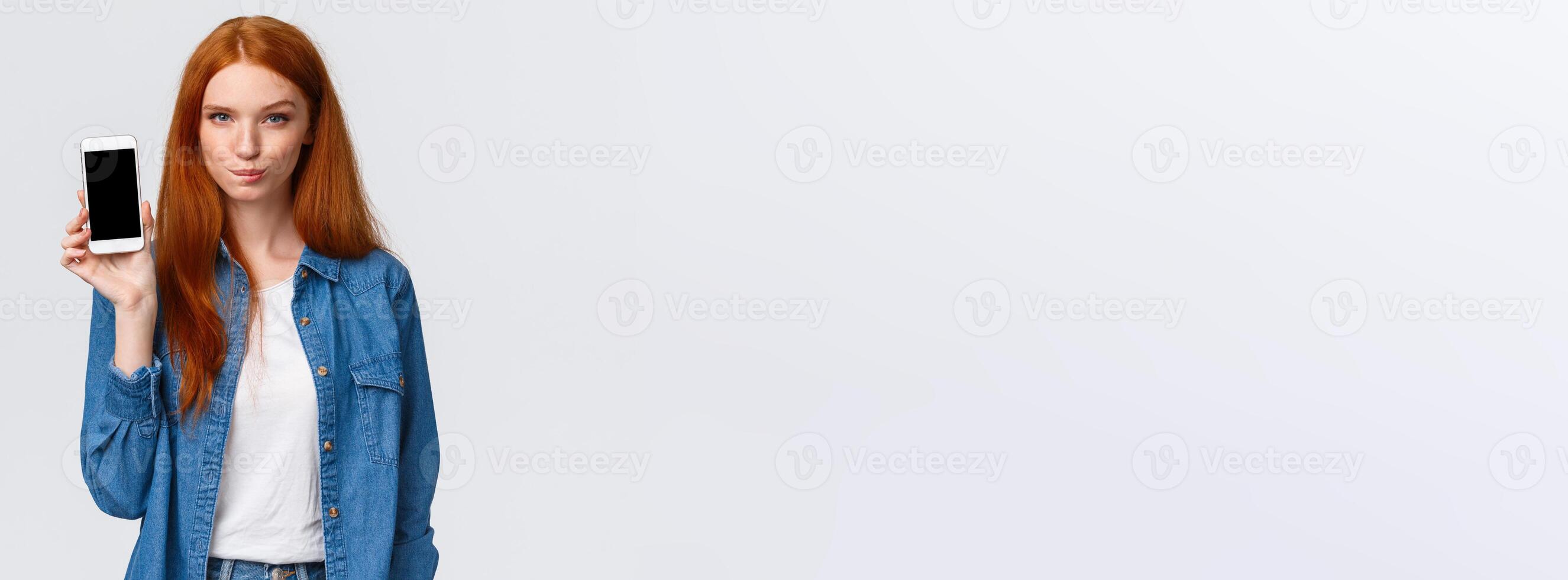 Sassy and daring good-looking redhead woman having interesting plan, showing her wishlist so know what buy on smartphone display, lick lips tempting and wishful, standing white background photo