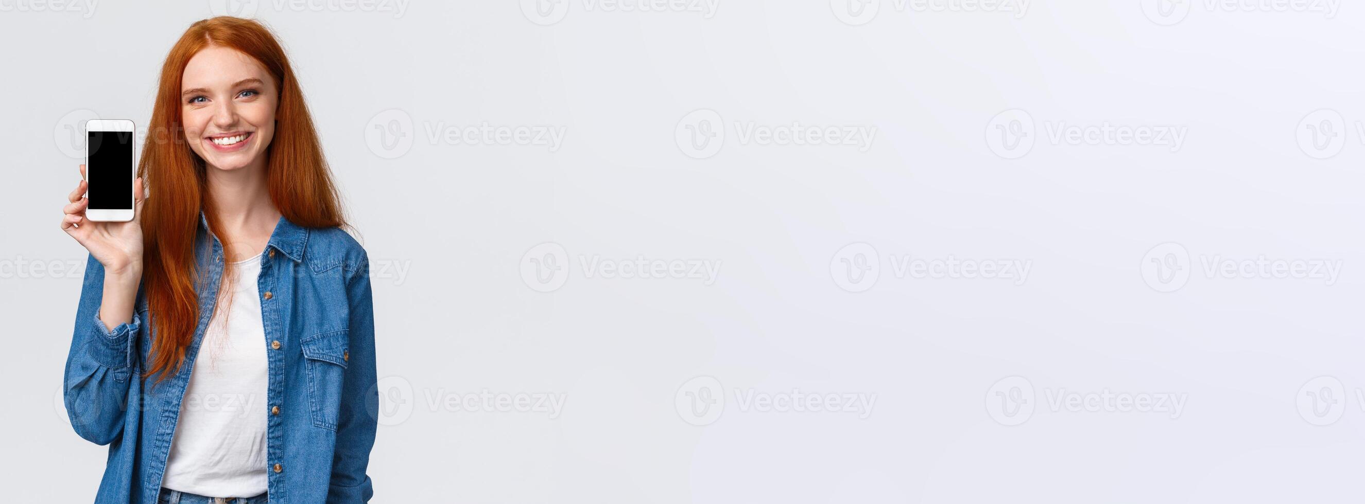 Cheerful friendly redhead woman checking water app, stay hydrated with smartphone application, showing mobile screen and smiling delighted, standing white background in casual clothes photo