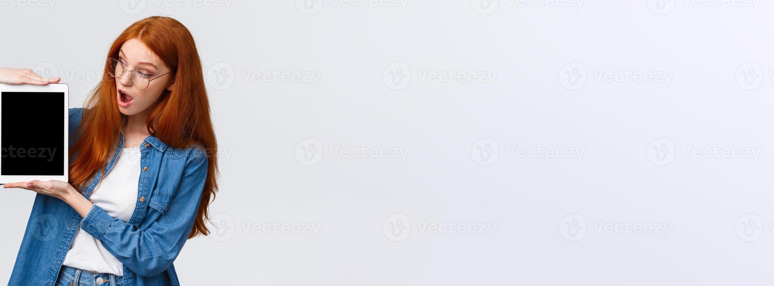 Surprised and curious cute redhead female in denim shirt, looking astonished digital tablet screen, say wow, checking out something awesome, standing white background photo