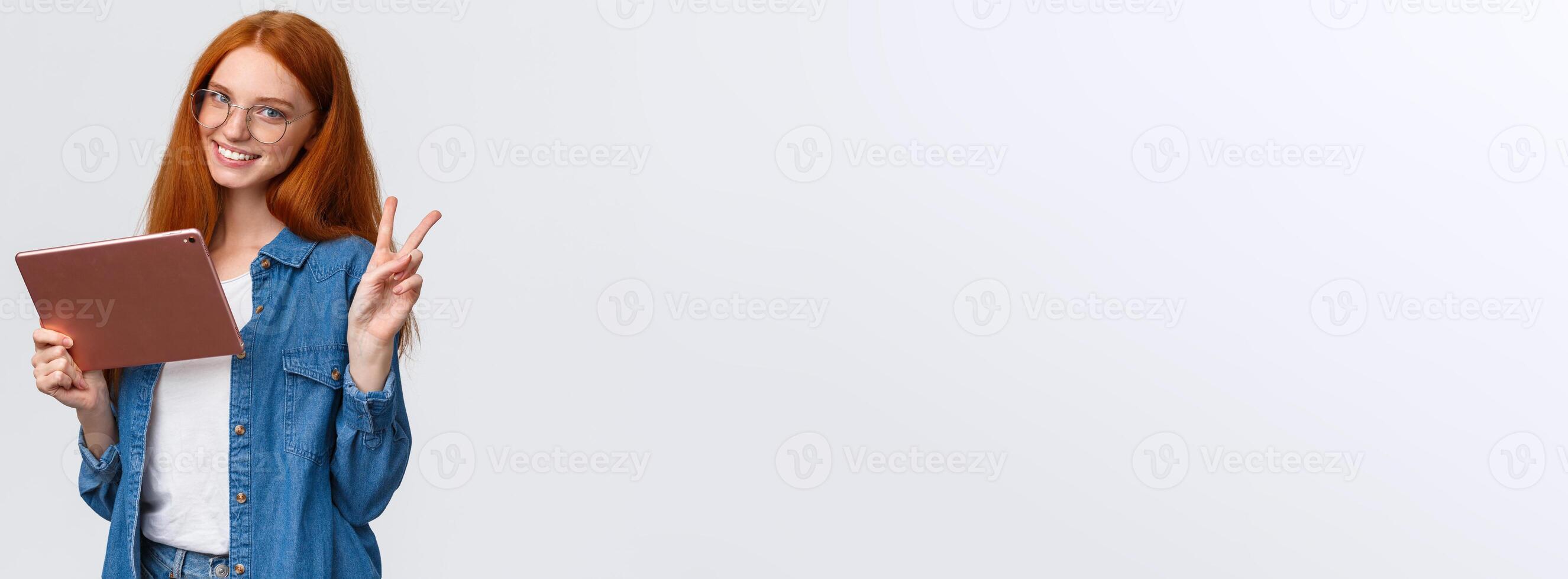 People, work and education concept. Cheerful attractive redhead female student in glasses, studying remote online courses, watching on digital tablet, showing peace sign and smiling photo