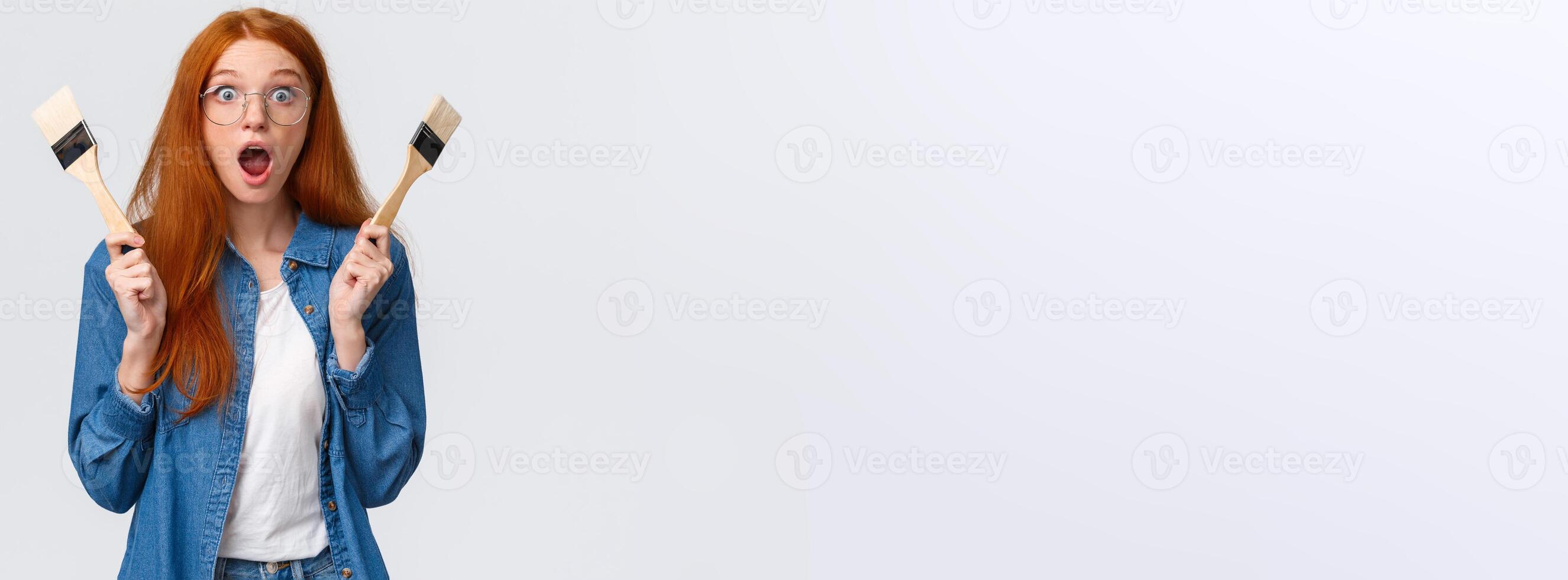 Amused and excited, wondered redhead woman creating new art, painting, look stunned and thrilled, say wow drop jaw and pop eyes camera, holding two paintbrushes, white background photo