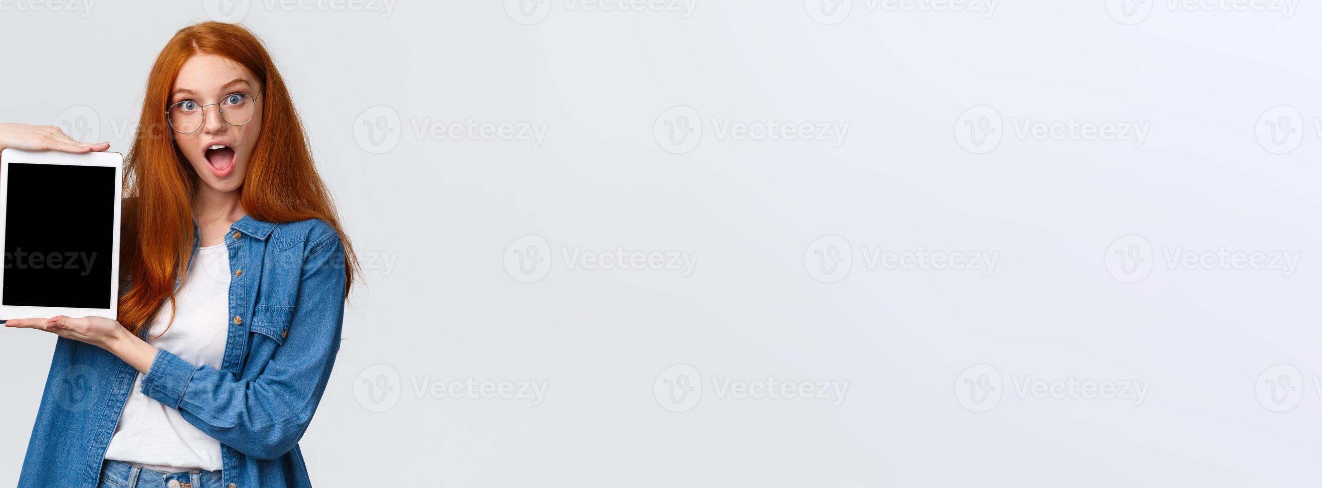 Excited and amused attractive redhead teenage girl making design, creative project on device screen, showing digital tablet display, look amazed, open mouth fascinated, white background photo
