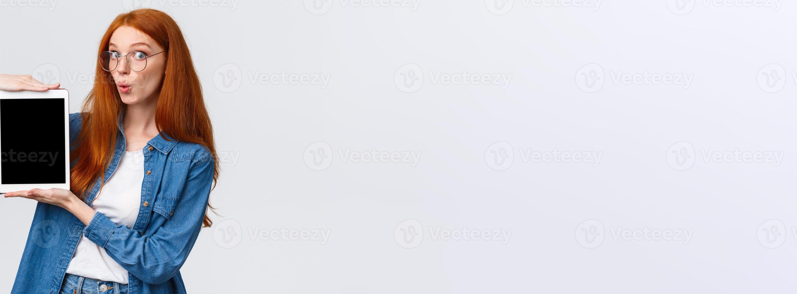 Amused, excited good-looking redhead woman in denim shirt, folding lips in wow sound, looking amazed camera, holding digital tablet, showing device screen, standing white background photo