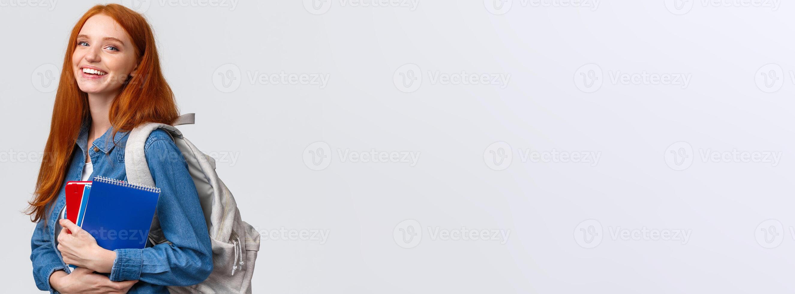 Teenagers, students and education concept. Cheerful lovely redhead female studying, going to univeristy or college, smiling carefree holding backpack and notebooks, standing white background photo
