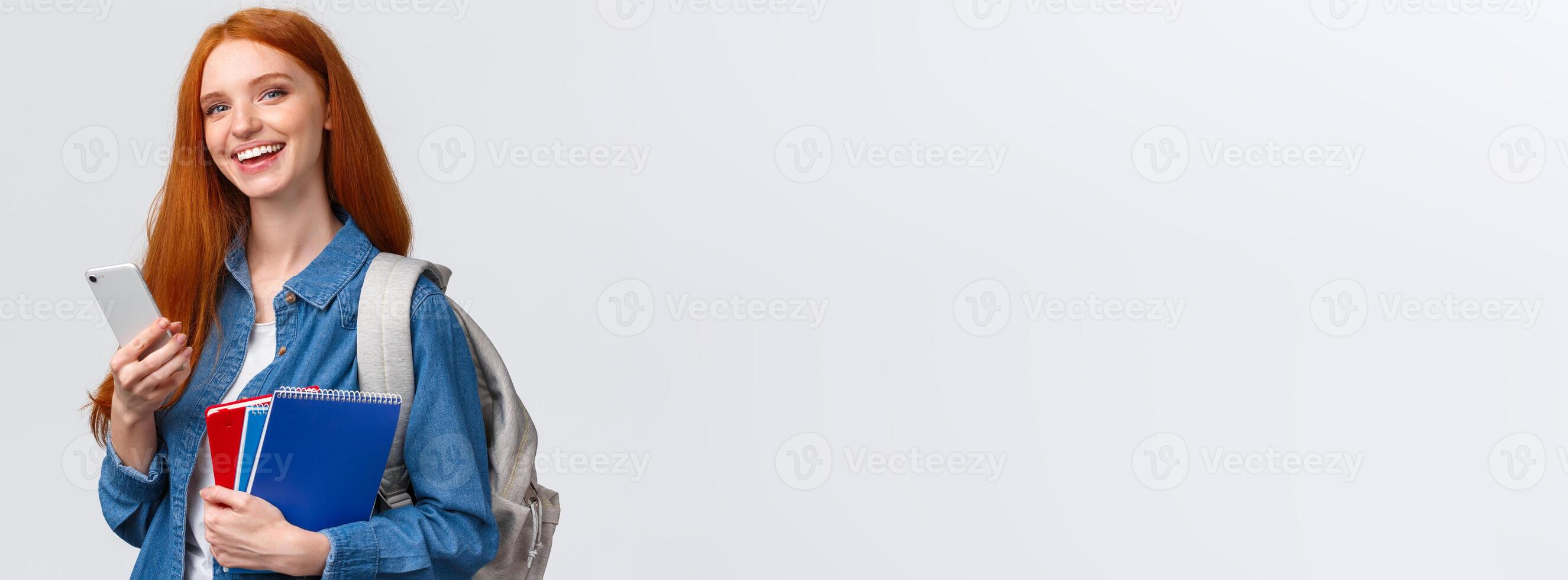 Charismatic, happy smiling redhead teenage girl, college student with backpack, notebooks, holding smartphone, texting friend, chatting or order taxi, waiting for lift grinning camera photo