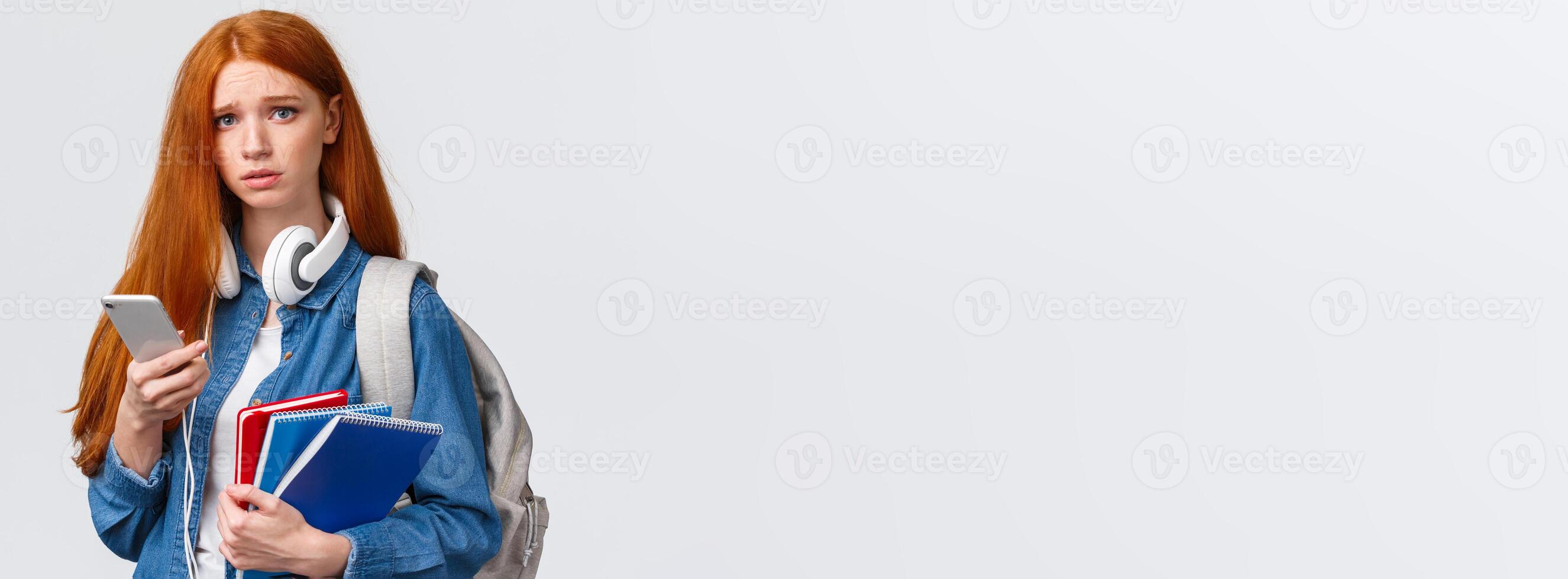 Uneasy, upset and gloomy cute redhead teenage girl, student in university frowning look distressed and unhappy camera, holding backpack education notebooks and smartphone photo