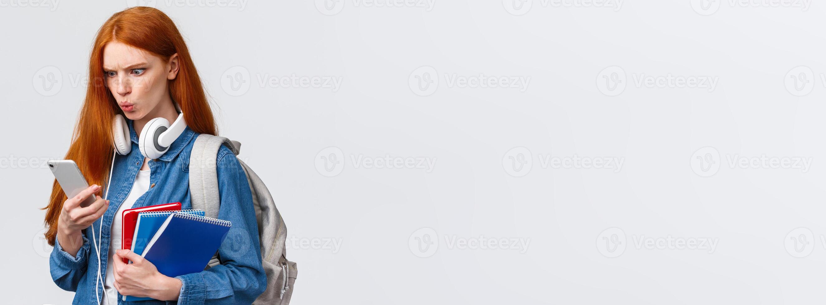 Displeased, mad or confused good-looking redhead college girl, student with backpack, notebooks and headphones, reading strange message on smartphone, frowning upset, white background photo