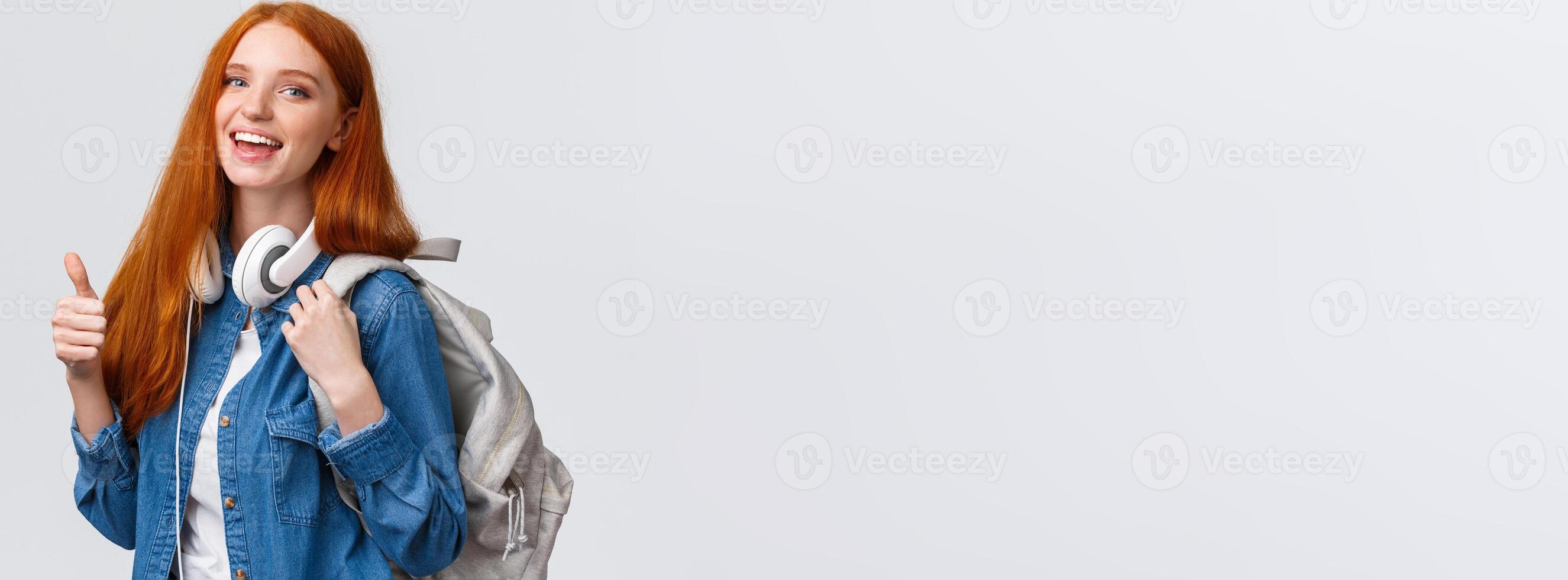 Waist-up portrait joyful cute redhead girl inviting freshmen apply univeristy, got scholarship, smiling showing thumbs-up in approval, recommendation, holding backpack and headphones photo