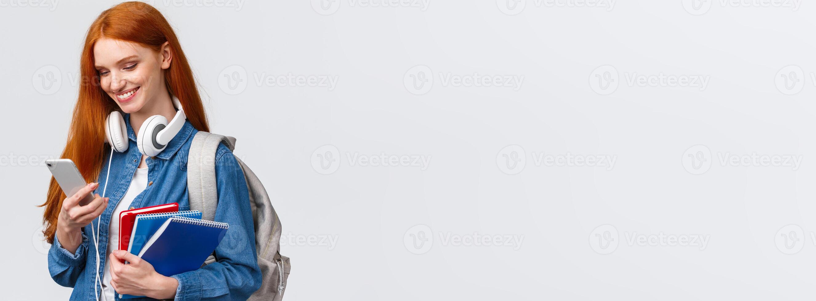 Teenagers, college and education concept. Attractive redhead female, univeristy student with headphones, backpack and notebooks, answer girlfriend message, queer love in air photo