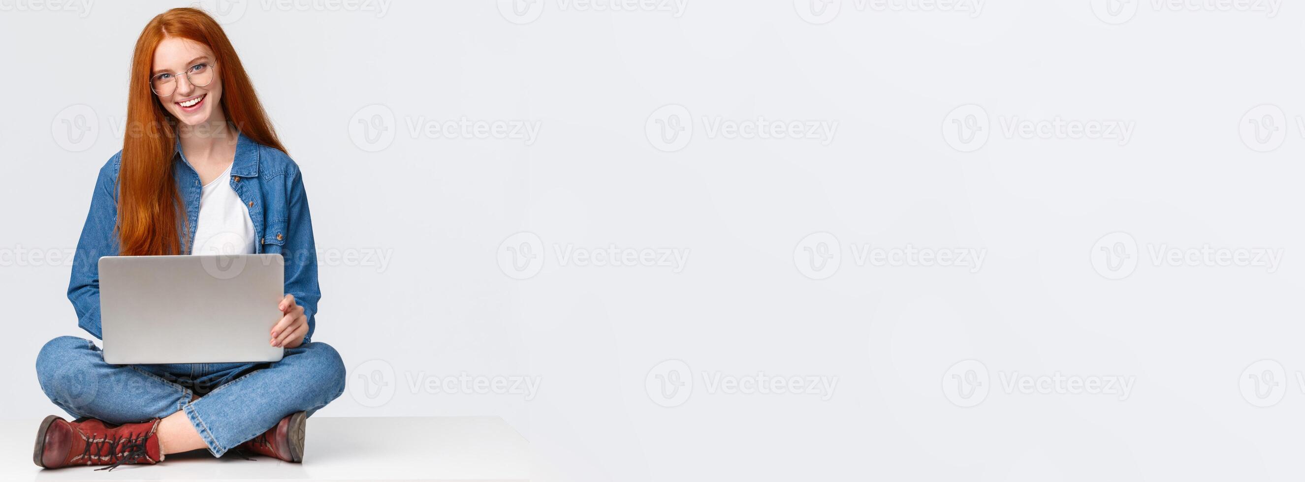 Cheerful sassy redhead female student, college girl in glasses, sitting on crossed legs with laptop, working freelance, prepare project awat from office, remote over white background photo