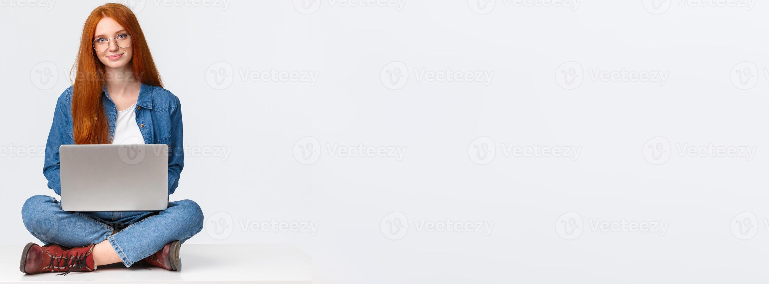 Education, modern lifestyle and college concept. Attractive carefree redhead female student sitting with legs crossed on floor, using laptop, working at coworking space, white background photo