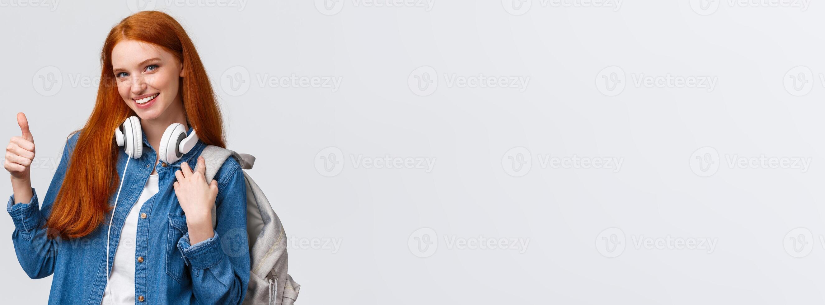 Girl found excellent part-time job after university classes. Attractive redhead woman with backpack, headphones over neck, showing thumb-up and smiling in approval, recommending photo