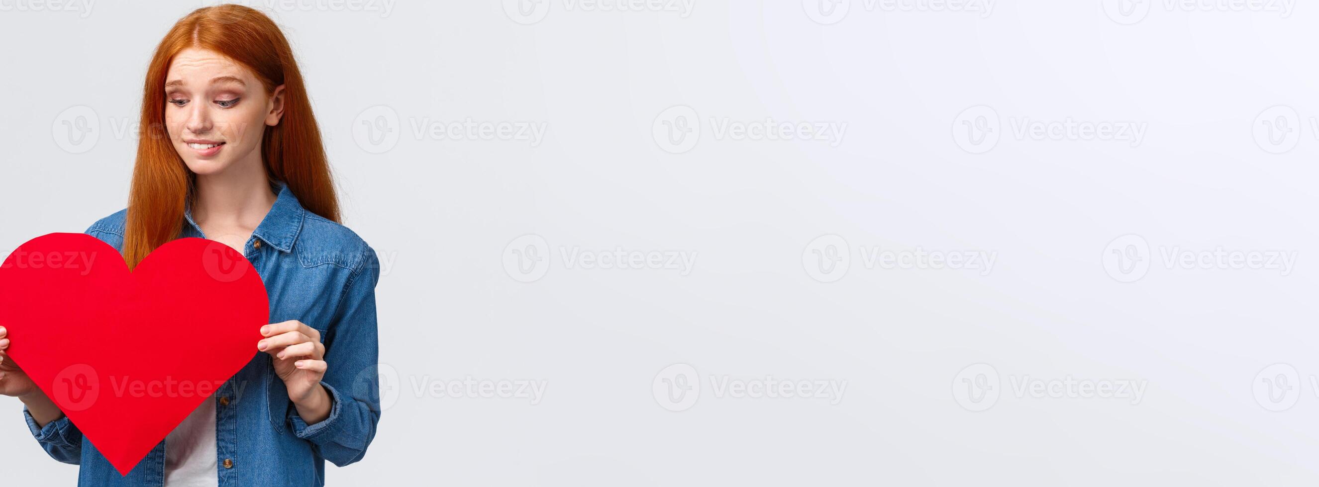 Indecisive cute and timid, shy redhead girl wants confess in love, show sympathy unsure give present on valentines day or not, holding big red heart, biting lip worried, white background photo