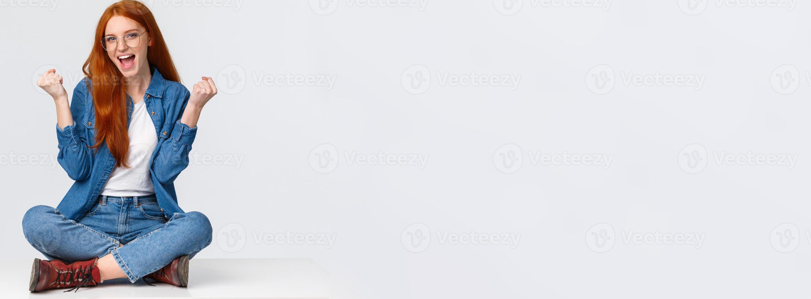 Cute and confident sassy redhead girl won challenge, sit floor, fist pump saying yes celebrating great news, winning competition, posing over white background satisfied, triumphing over achievement photo