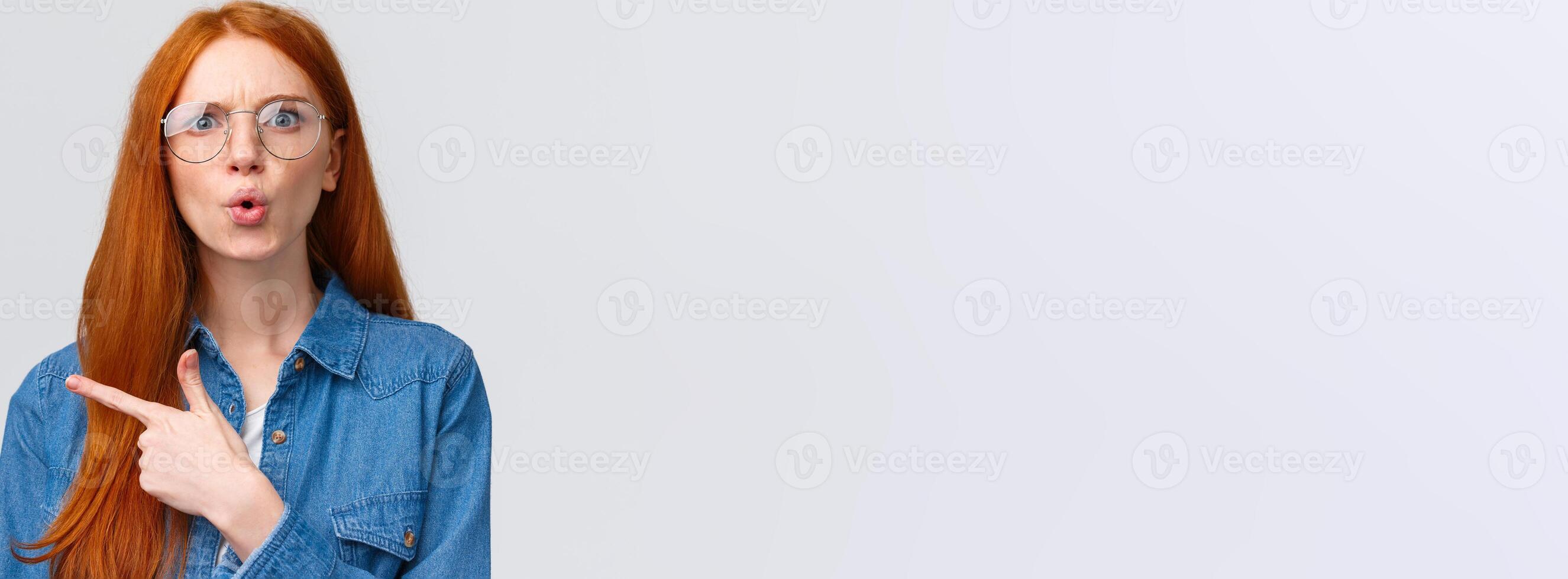 Confused and shook questioned redhead woman asking question and stare shocked and displeased, pointing finger left at something bothering and strange, standing complaining over white background photo