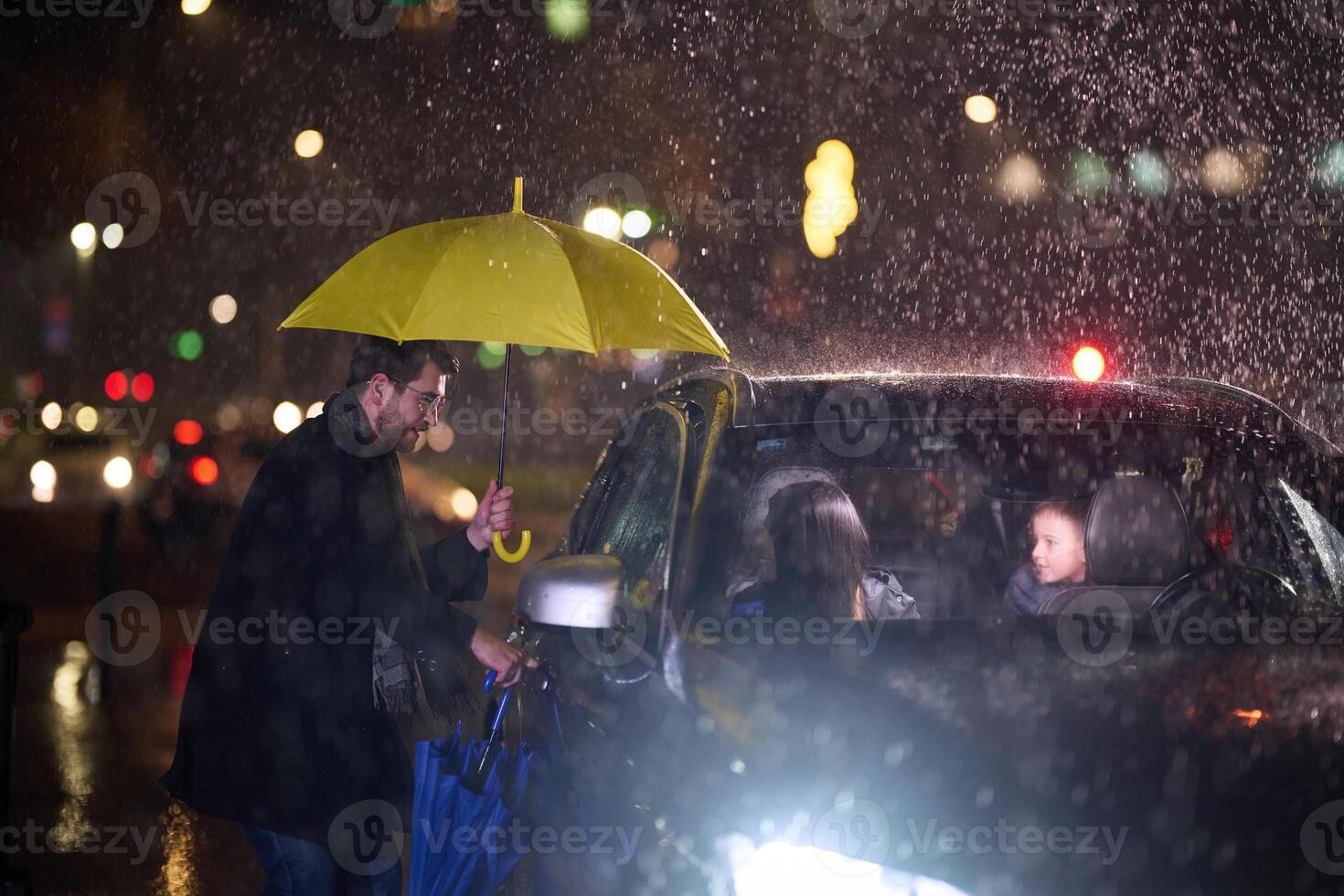 A family man lovingly opens the car for his family in rainy weather, sheltering them with an umbrella photo