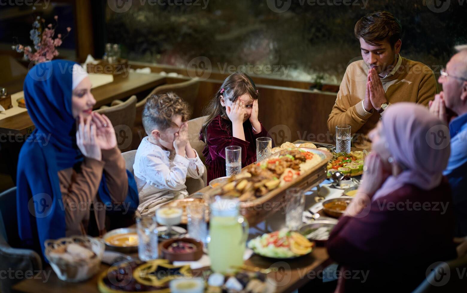 In a modern restaurant setting, a European Islamic family comes together for iftar during Ramadan, engaging in prayer before the meal, uniting tradition and contemporary practices in a celebration of photo