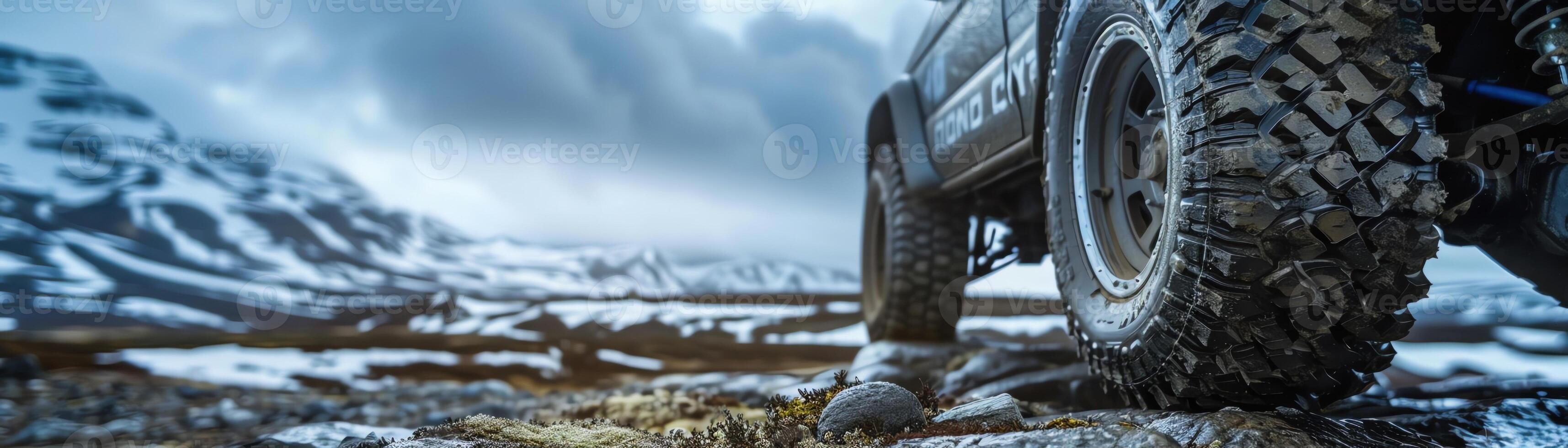 Rugged offroad vehicle tire on a rocky terrain, blurred snowy mountain background, dramatic overcast sky photo