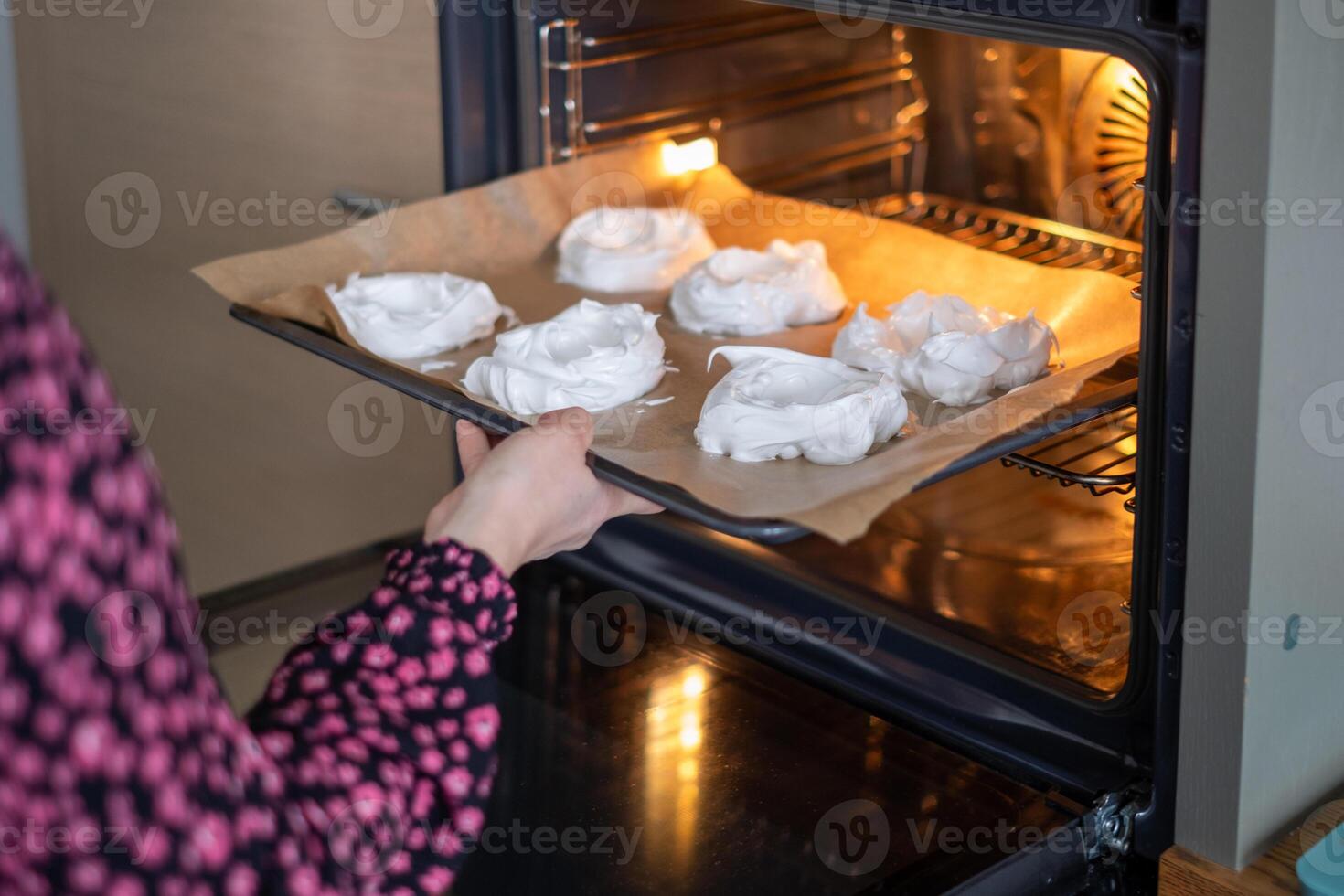 A person in a patterned shirt slides a tray of meringues into the oven, an intimate moment in home baking, perfect for illustrating recipe steps or baking classes. photo