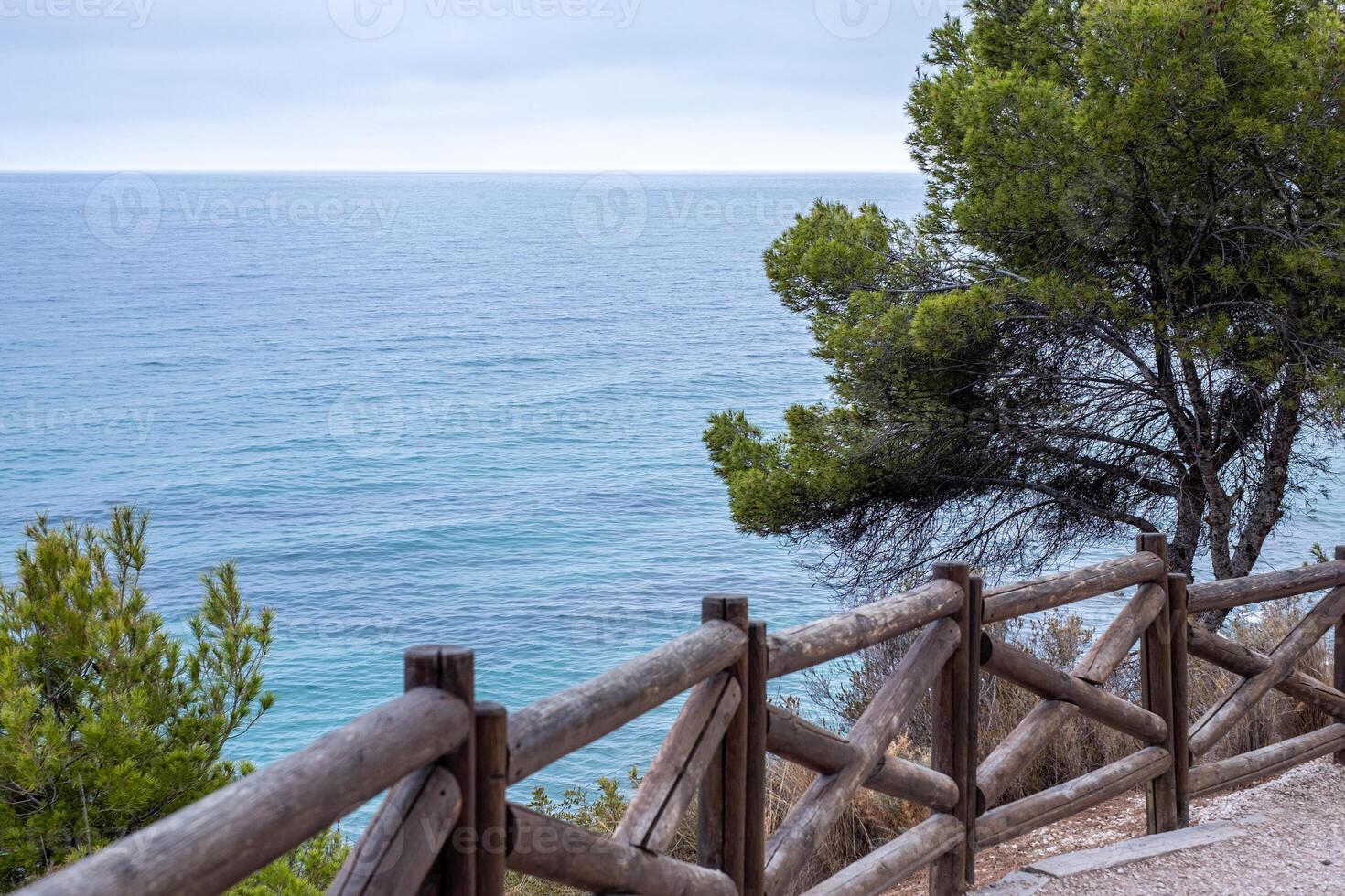 Coastal view from a rustic wooden fence, greenery on the side, overcast skies above, suggesting tranquility or travel themes. photo