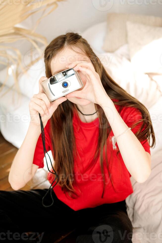 Young woman with long brown hair in a red T-shirt takes pictures with a film camera while sitting on the floor at home. Portrait of a young woman with a camera in front of her face photo