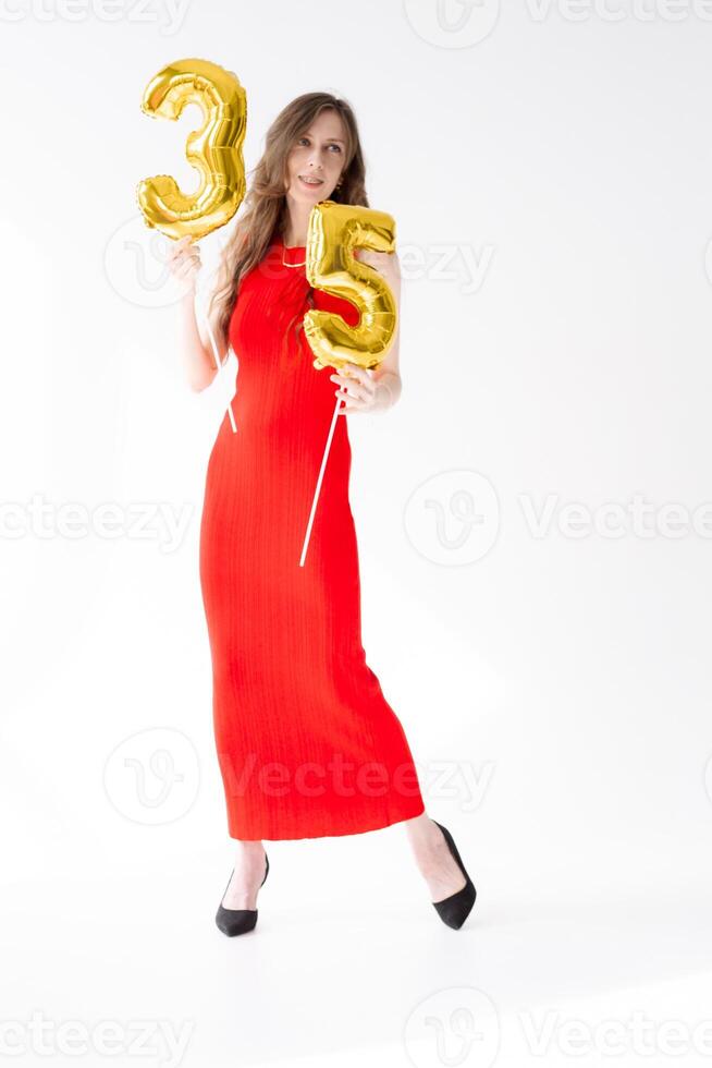 A beautiful woman with balloons and the numbers 35 is photographed in the studio in a red dress and shoes. Beautiful and feminine woman celebrates her 35th birthday photo