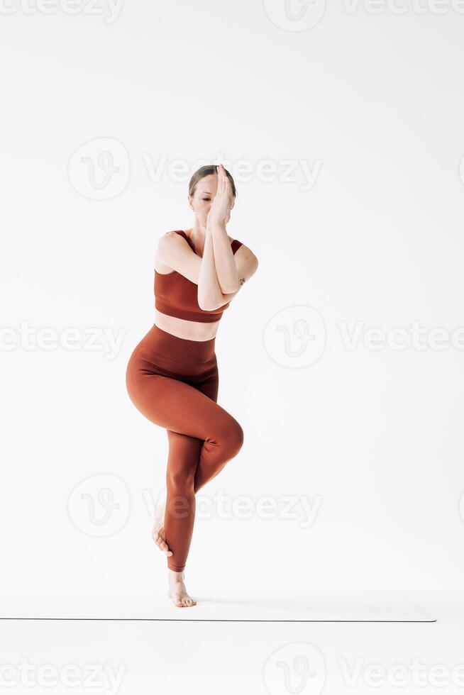 Slender woman in a brown sports uniform stands in garudasana asana. Yoga classes on white background photo
