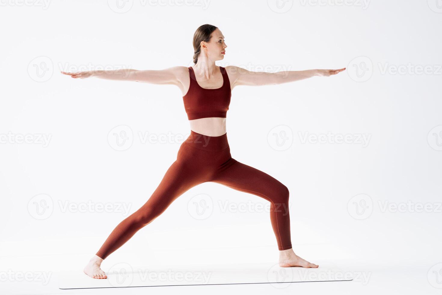 Young and tall woman does Virabhadrasana on a mat against the background of a white wall. Yoga training indoors photo