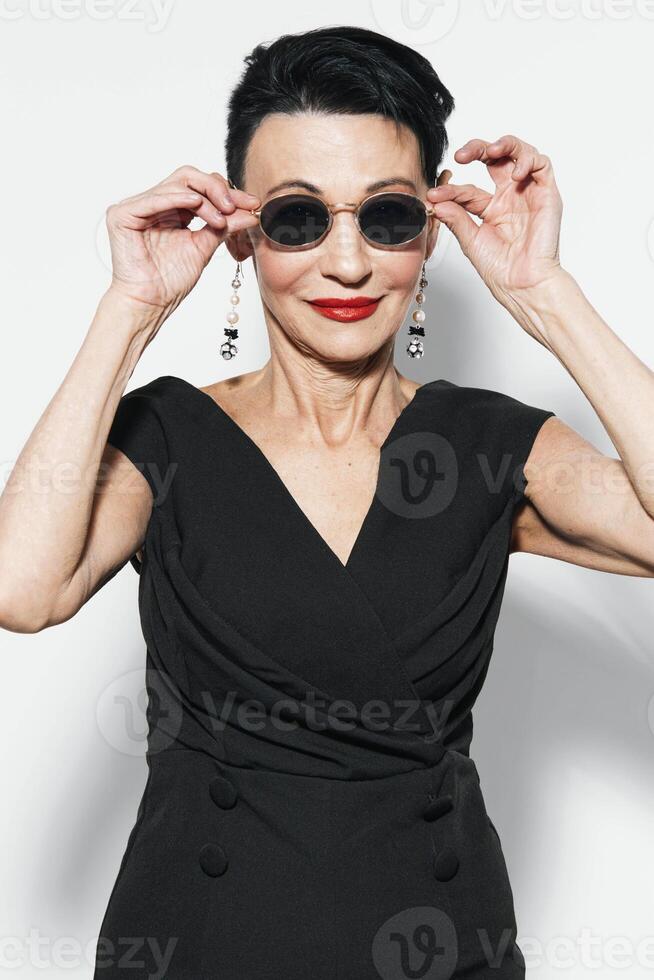 Elegant older woman in black dress and sunglasses covering her face with hands in dramatic pose photo
