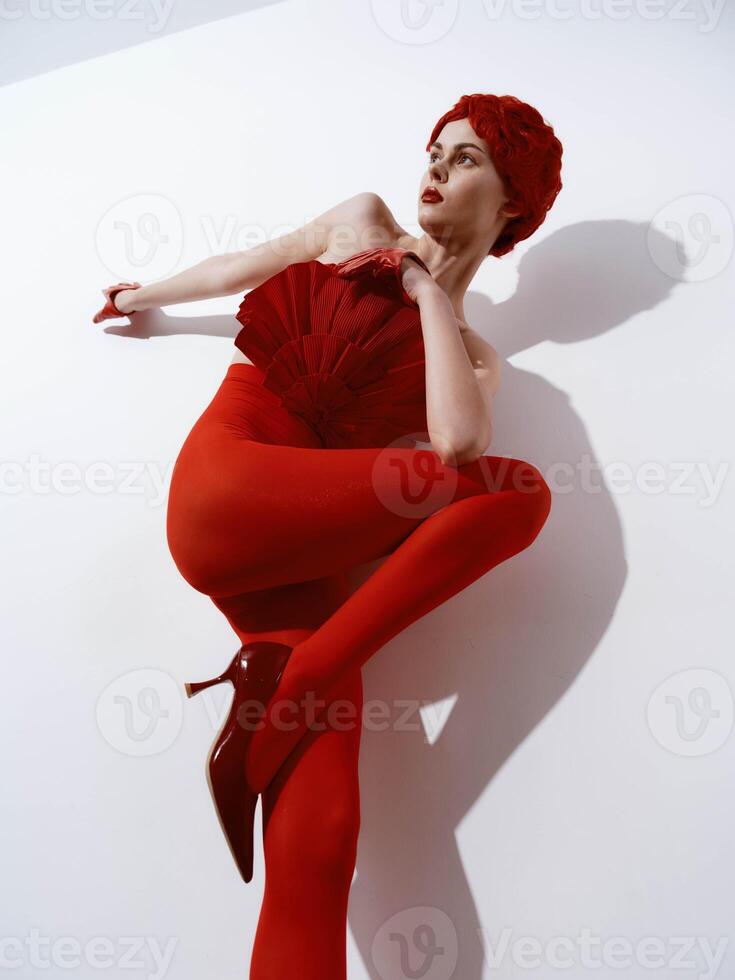 Stylish woman in a red dress and high heels posing against a white wall with a shadow photo