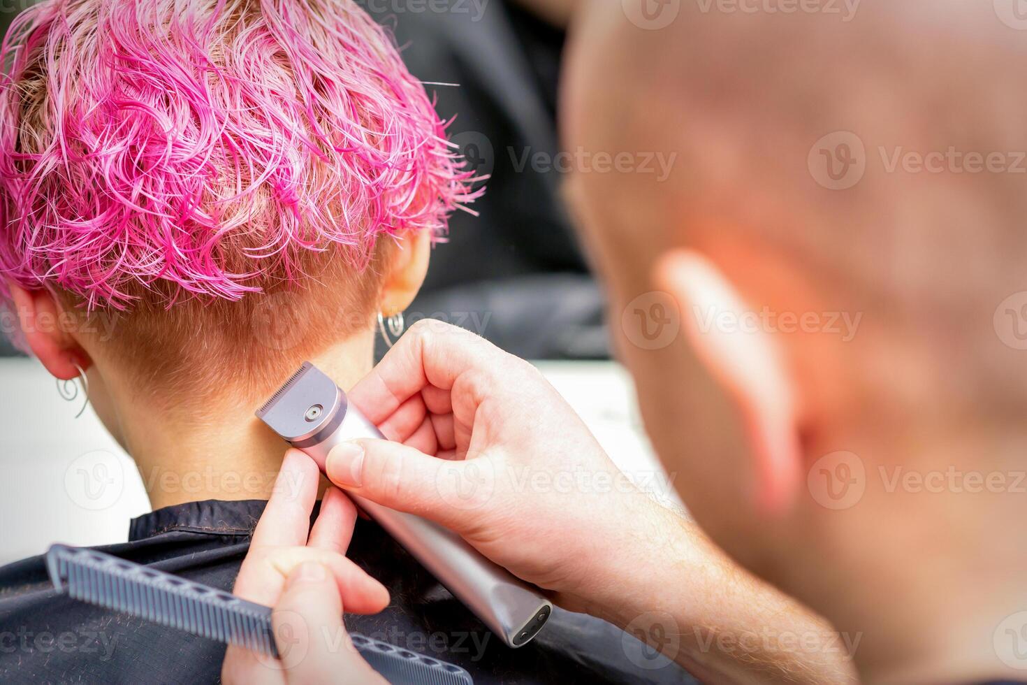 Hairdresser shaving nape and neck with electric trimmer of a young caucasian woman with short pink hair in a beauty salon. photo