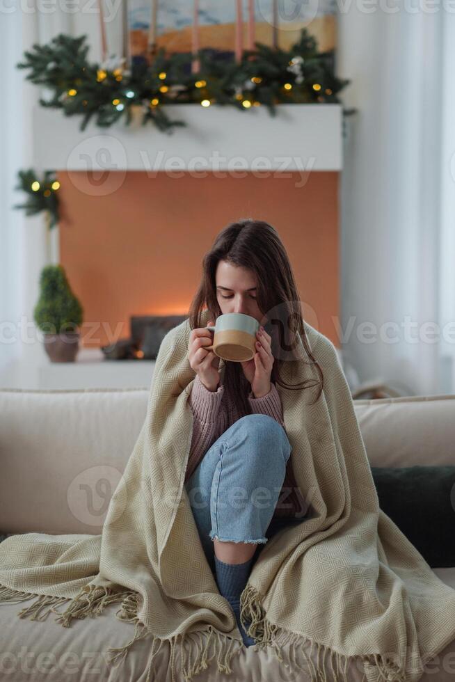 Woman freezes in winter time. Young girl wearing warm woolen socks and wrapped into plaid, holding a cup of hot drink while sitting on sofa at home. Keep warm. photo