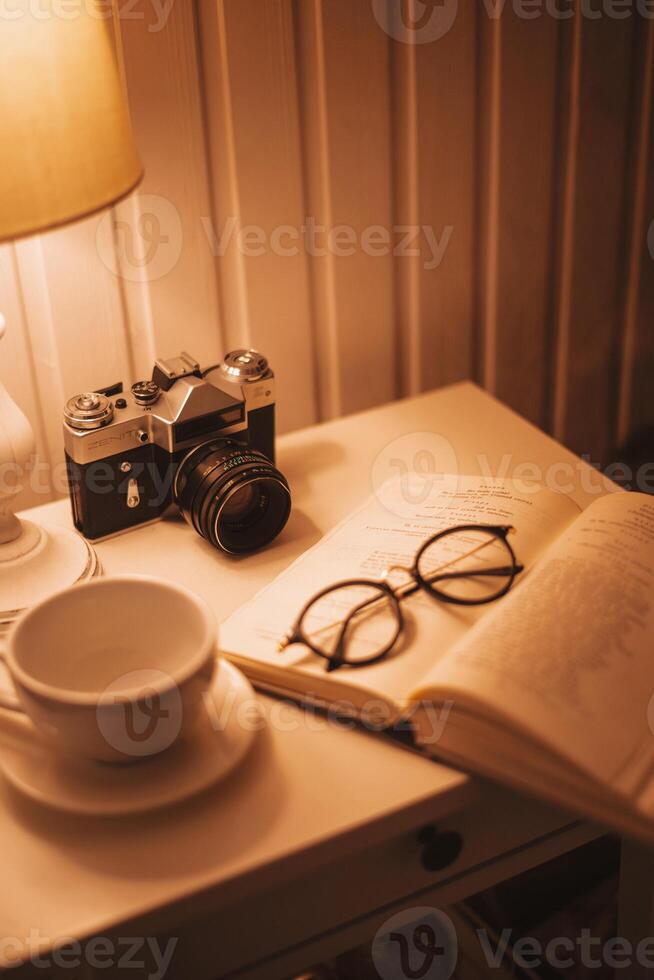 November 28, 2020. Kyiv, Ukraine. Vintage home atmosphere. Retro photo camera, glasses, coffee cup and book on the table with lamp. Selective focus