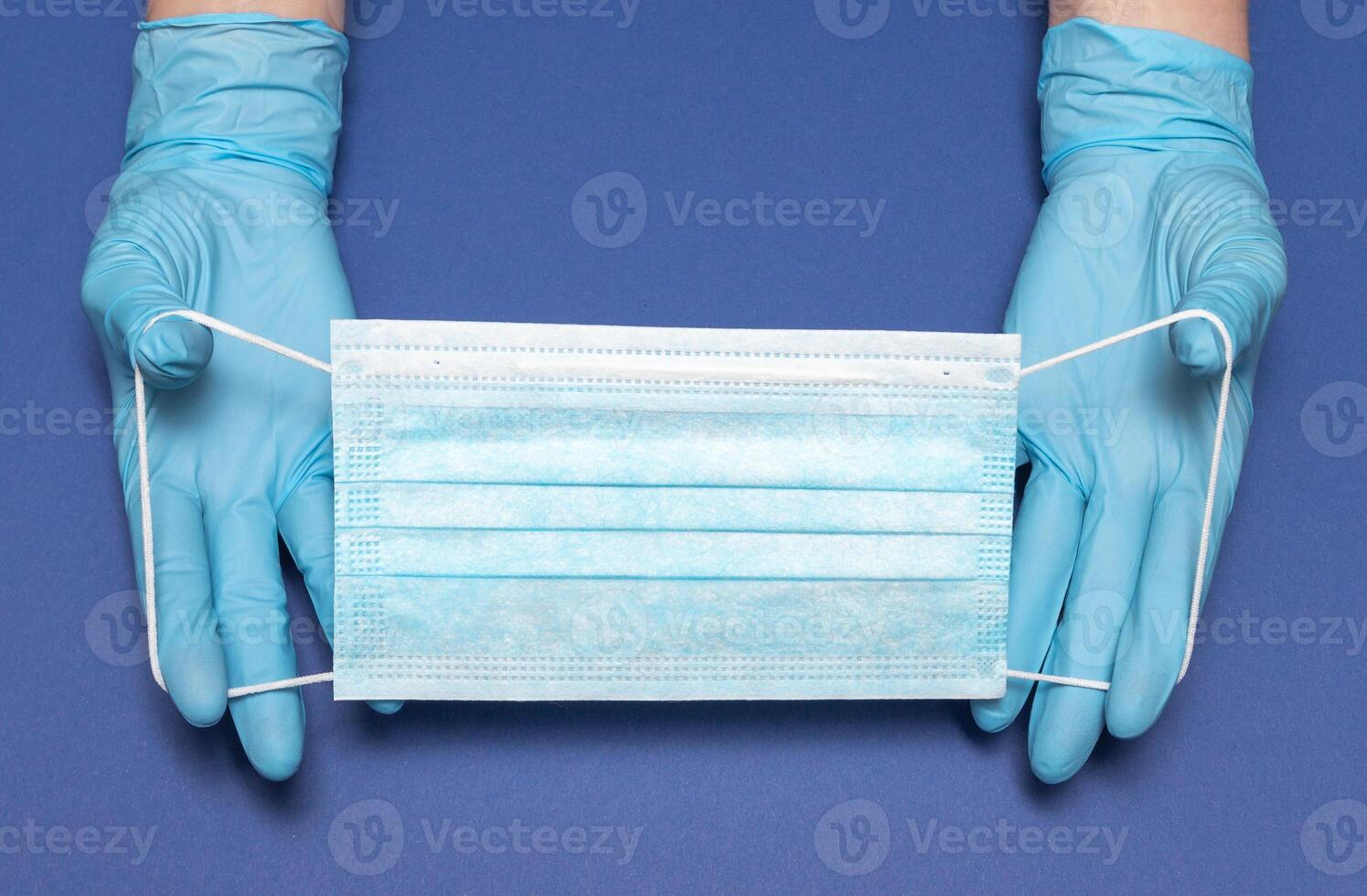 Hands in blue medical gloves is holding face mask on blue background. Protective respiratory medical mask against spreading virus. Close up, copy space, top view, flat lay. photo