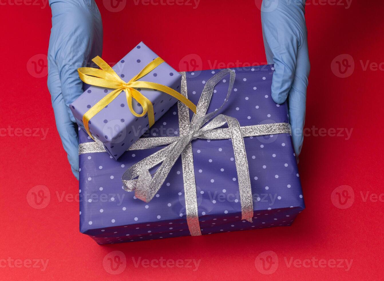 Hands in blue latex gloves holds gift boxes in color paper with polka dots with tied ribbon with as a present for Christmas, new year, valentine day or anniversary on red background. Top view photo