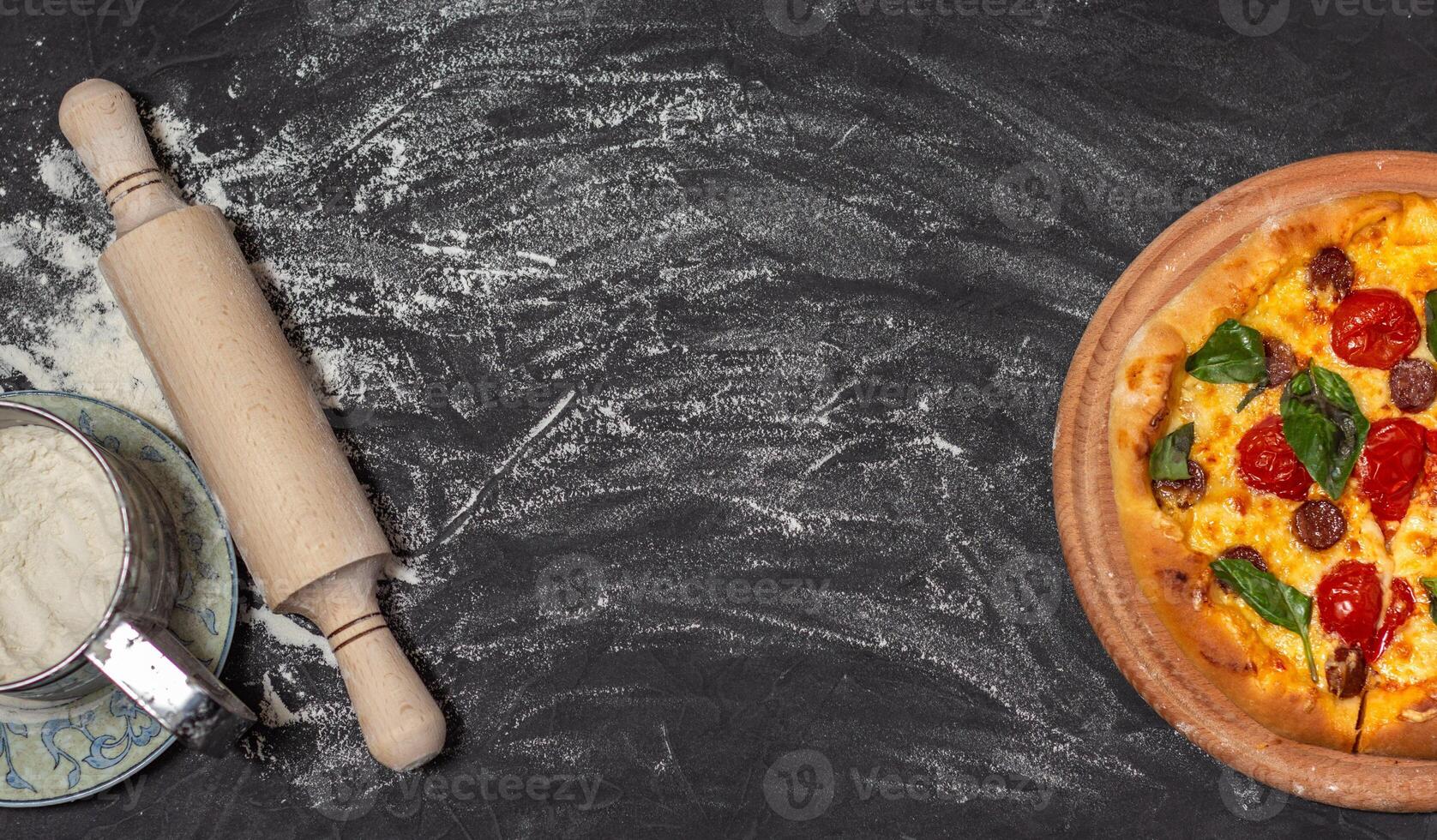 Pizza on wooden plate on black stone background. Rolling-pin and floor. Cheese pizza with tomato, mozzarella and basil. Banner. Close-up photo. Copy space. photo