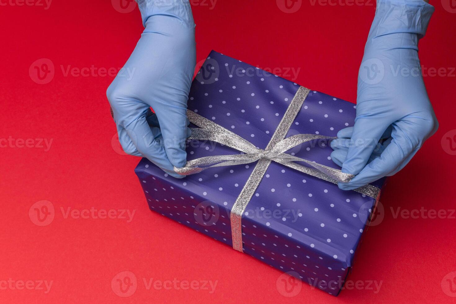 Hands in blue latex gloves holds gift box in color paper with polka dots with tied ribbon with as a present for Christmas, new year, valentine day or anniversary on red background. Top view photo