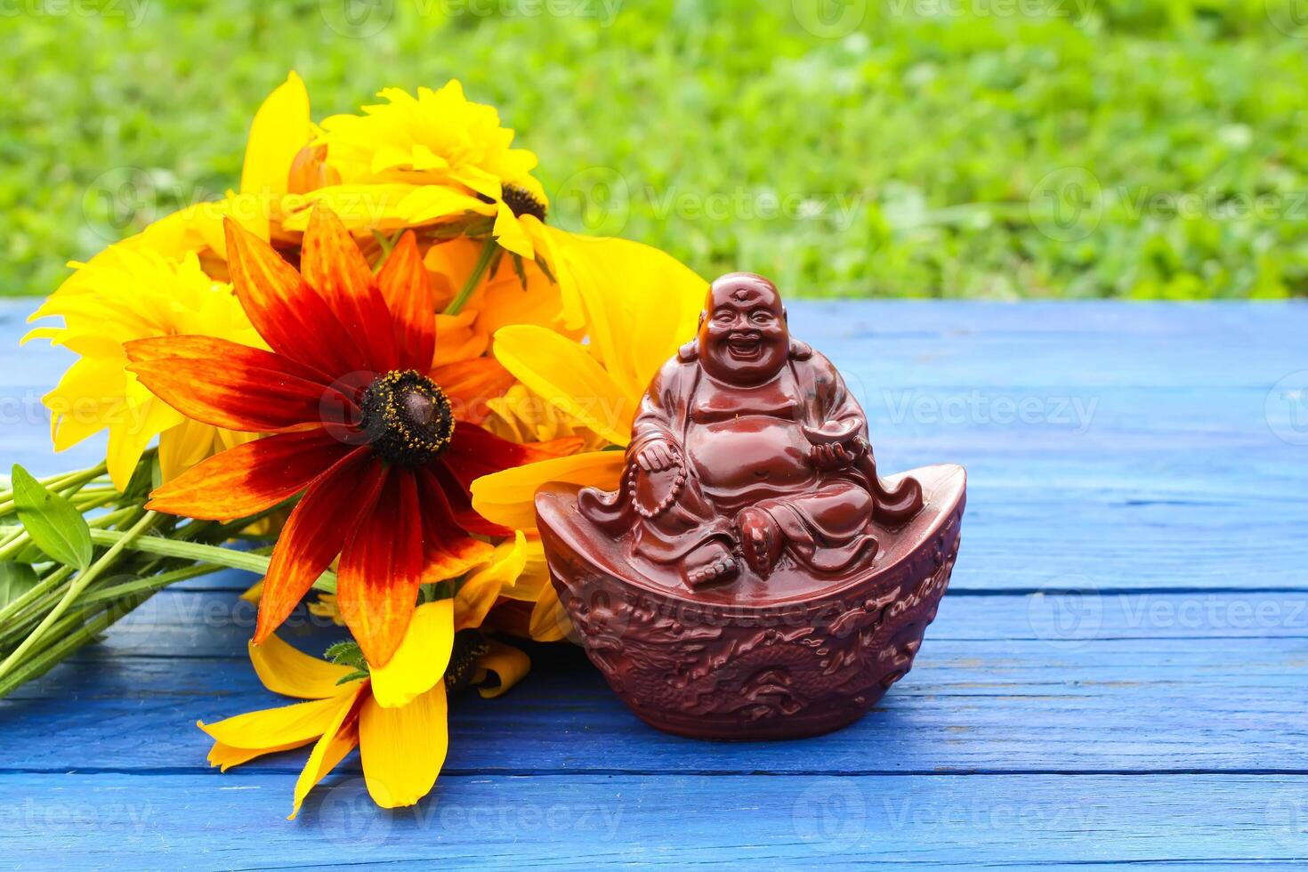 Ceramic buddha figurine on blue aged wooden boards in summer park with flowers. photo