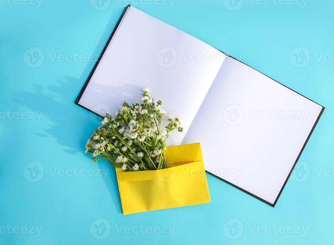 Yellow paper envelope with small garden white chamomile flowers and empty notepad on light blue background. Festive floral template. Greeting card design. Top view. photo
