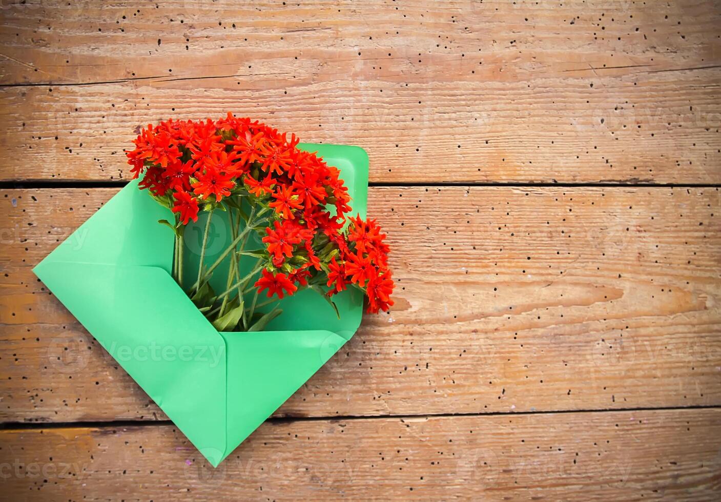 Green paper envelope with fresh garden red lychnis flowers on wooden background. Festive floral template. Greeting card design. Top view. photo
