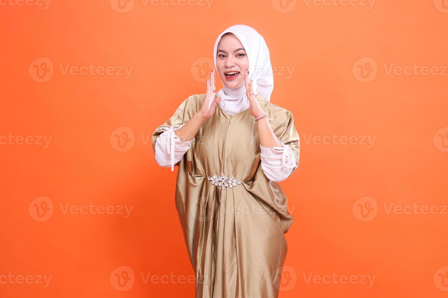 Cheerful beautiful young Asian woman model both hands announcing wearing Muslim dress kaftan with hijab, isolated on orange background. Concept of lifestyle, stylish fashion, promotion photo