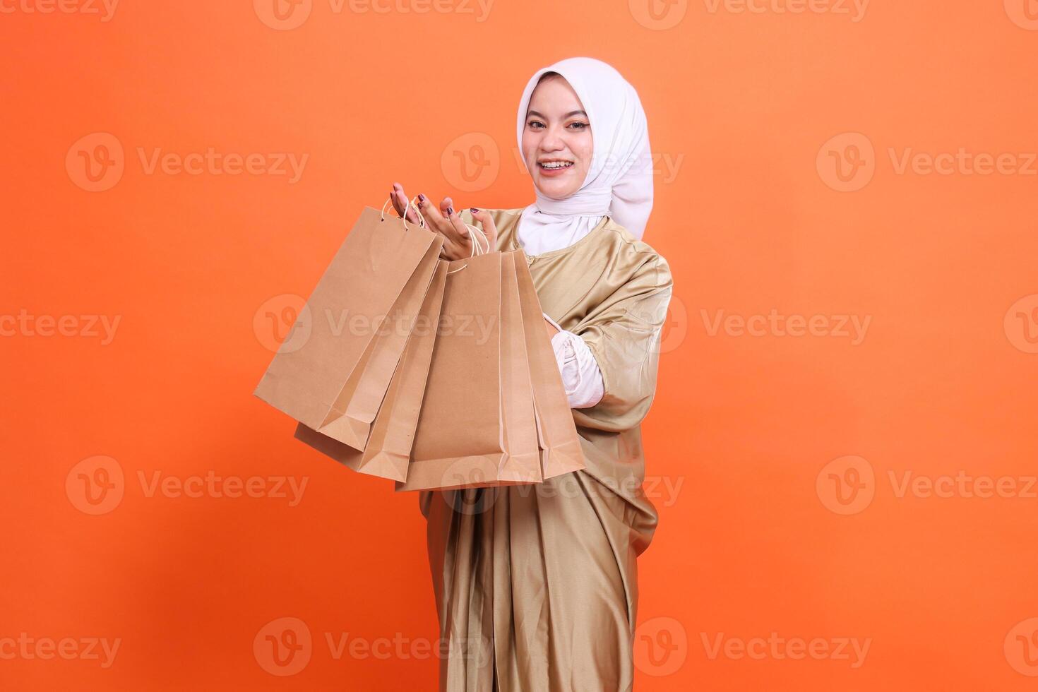 Cheerful Asian woman with both hands carrying a brown paper bag containing groceries in front of her wearing a kaftan Muslim dress with a hijab, isolated on an orange background. fashion transaction photo