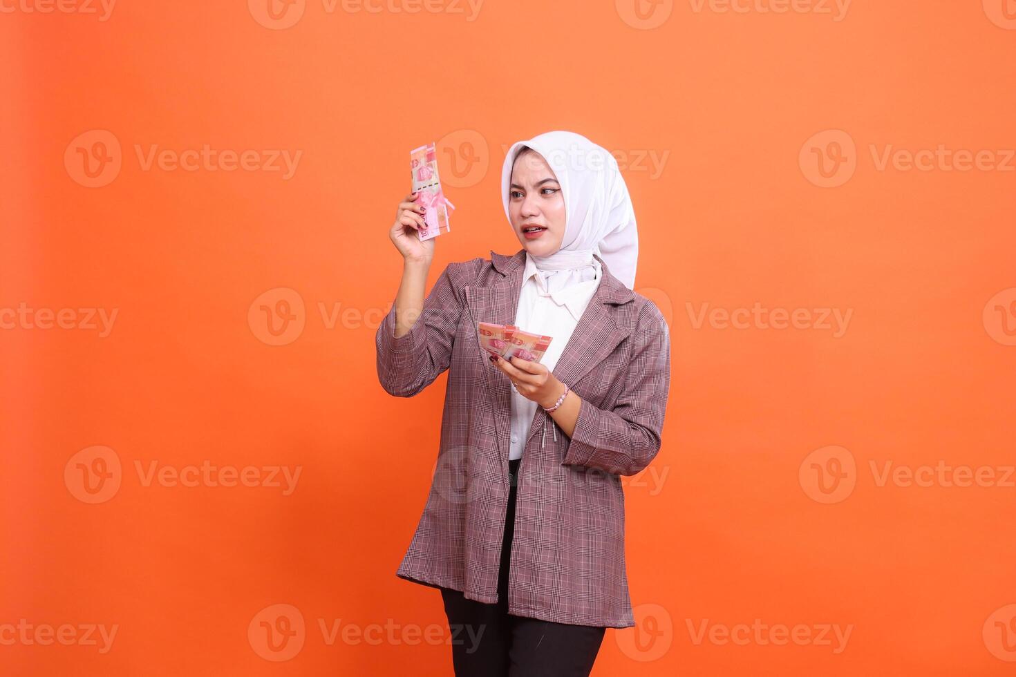 charm of beautiful indonesia woman wearing hijab shocked both hands holding and counting rupiah banknotes candid isolated on orange background. finance, lifestyle, advertising concept photo