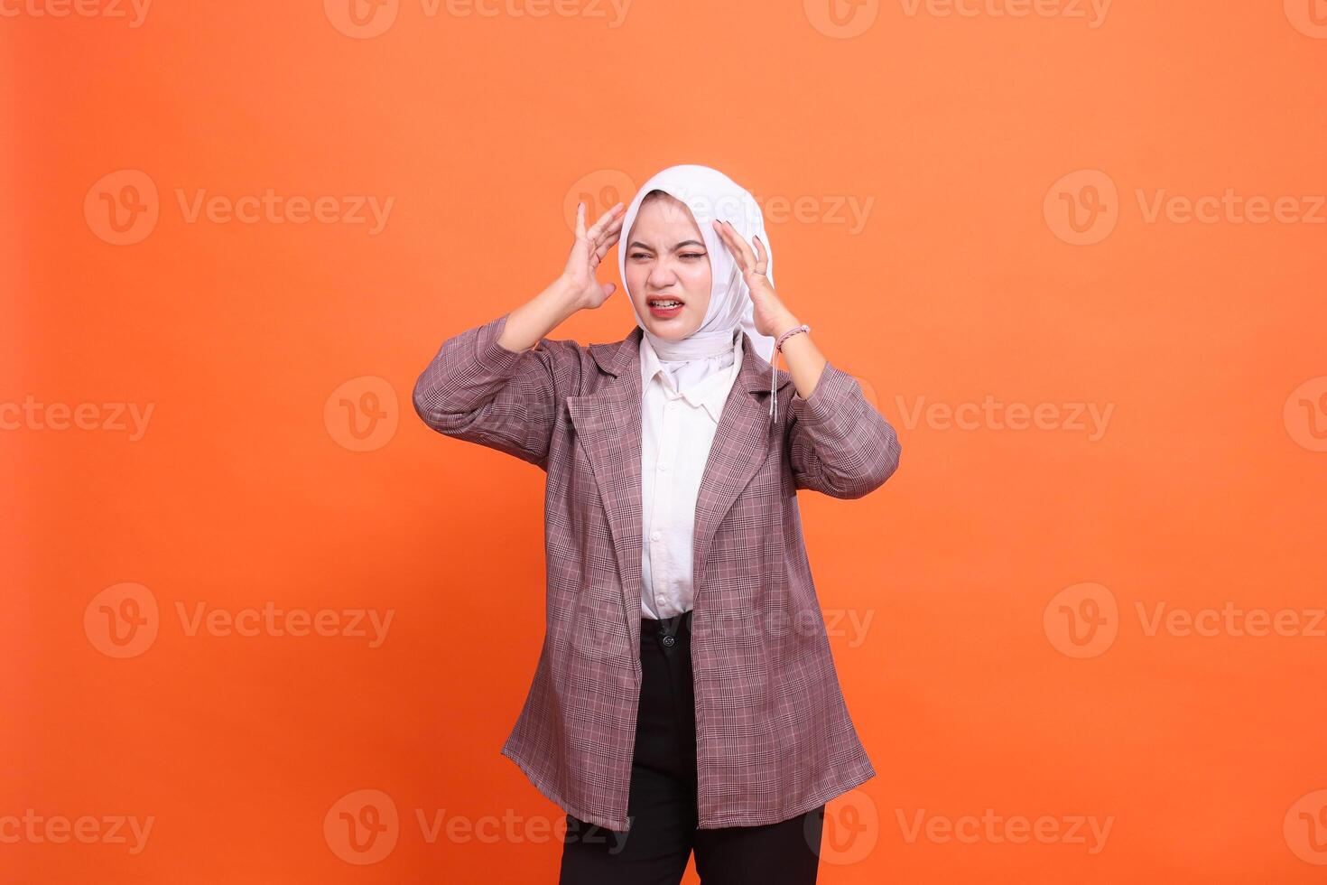 beautiful young Asian woman wearing a hijab grimacing in candid pain, experiencing pain, dizziness, position of both hands on her head with an orange studio background. health care, advertising photo