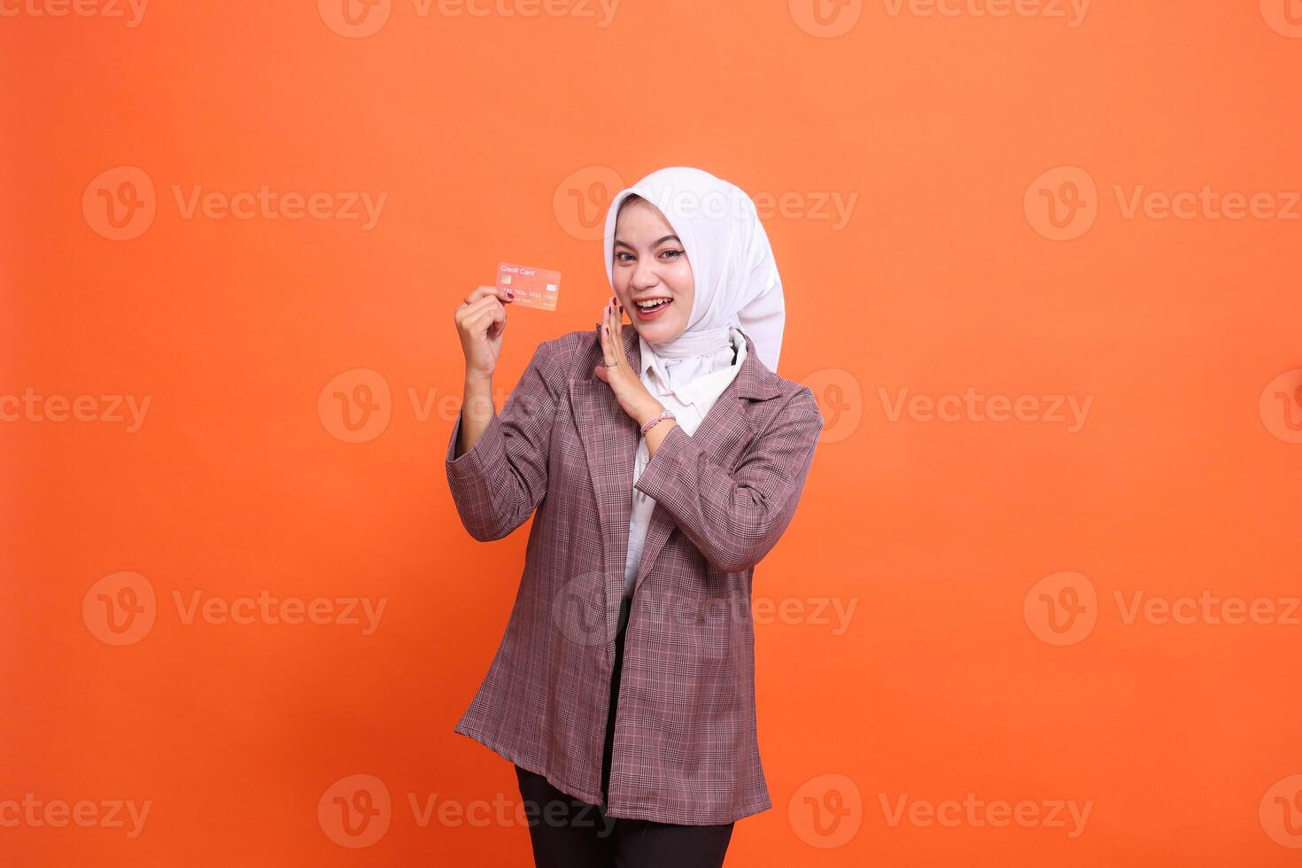 Young Asian woman cheerful hijab suit holding plastic debit credit card while hand whispering isolated on orange background. satisfied with service for financial concept, transaction, service photo