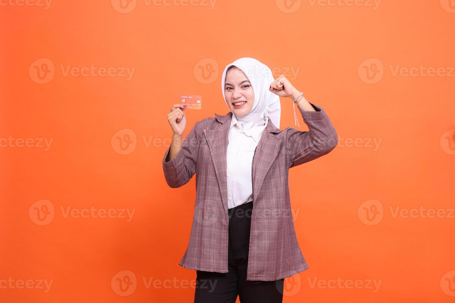 Cheerful young Asian woman in hijab suit holding plastic debit credit card while holding hands up isolated on orange background. satisfied with service for financial , mobile transactions, payments photo